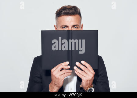 Professionalism. Confident businessman in classic wear covering his face with notebook and looking at camera while standing against grey background. Business look. Success concept Stock Photo