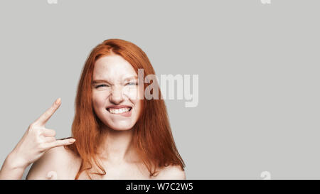 Studio shot young and cute teenage girl with long red hair showing hand sign and looking at camera while standing against grey background. Gesturing. Carefree Stock Photo