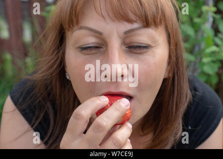 A young woman with red hair bites ripe strawberries and closes her eyes with pleasure. The concept of healthy eating and vegetarianism. Stock Photo