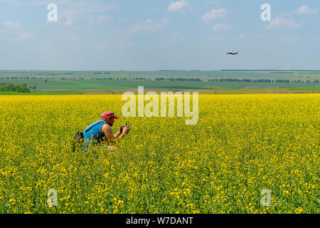 Russia, Kazan - June 23, 2019: A young man in a red cap and blue t-shirt launches a drone on a yellow field of flowers on a sunny day. The quadcopter Stock Photo