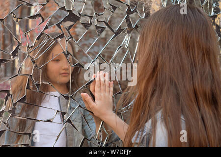 Young girl looks in a broken mirror and shows her hand on a mirror Stock Photo