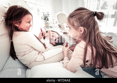 Cute sisters drawing on their dads face while he sleeps Stock Photo