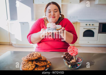 Fat young woman in kitchen sitting and eating sweet food. Plus size model hold small cakes in hands and look at them. Happy woman likes to eat. Body p Stock Photo