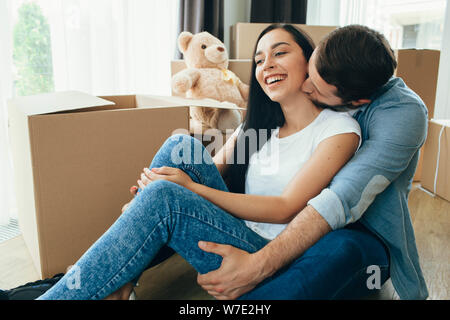 lovely man kissing his wife while sitting on floor at new home. Happy couple moving home, beginning new life to new apartment Stock Photo