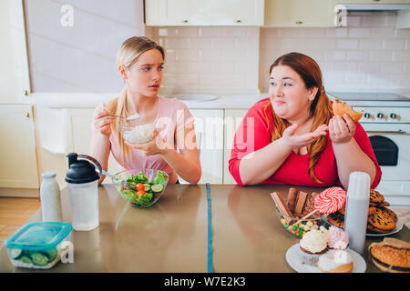 Confused young slim model eat farmer cheese at table. She look at plus size model who eats burgers and junk food. Happy body positive. Healthy and unh Stock Photo