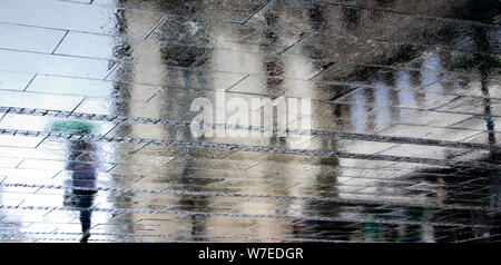 Blurry reflection shadow silhouettes of a woman walking alone under umbrella on a rainy pedestrian city wet street  on a summer day, in a puddle Stock Photo