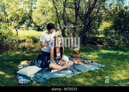 Young couple on a picnic sitting on a blanket The man put on her head flower crown Selective focus Stock Photo