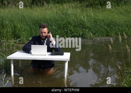 Handsome young man talking on the phone and using the computer while working. At the table, standing in the swamp, in the water Stock Photo