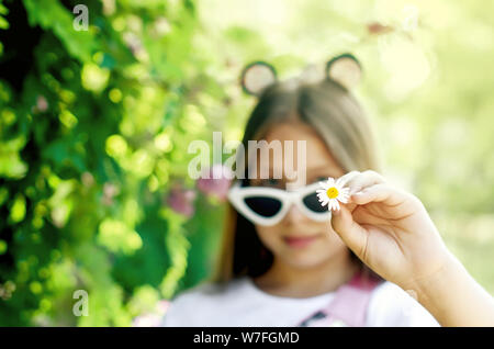 smiling and happy teen girl in funny white glasses with fluffy blond long hair holds a chamomile color in her hand on a summer sunny day near a green Stock Photo