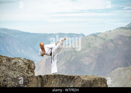 Karate athlete is standing on mountains cliff and demonstrating kicks against valley view Stock Photo