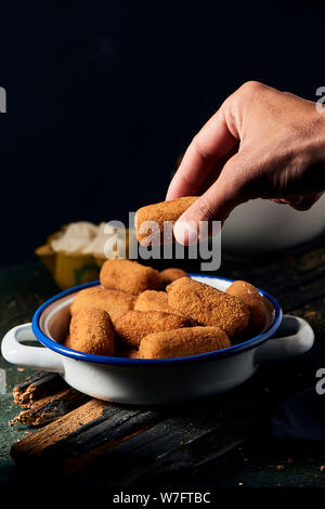 closeup of a man picking a spanish croquette from a white and blue enamel plate, placed on a rustic wooden table, and a small yellow mortar with aioli Stock Photo