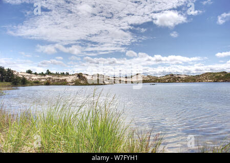 View of vegetated sand dunes across a body of water with tall grass in the foreground and a partly cloudy sky in the background Stock Photo