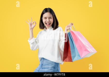 Cheerful beautiful Asian woman holding multi coloured shopping bags and shows ok sign on light yellow background. Stock Photo