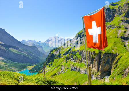 Waving flag of Switzerland on the hill in Swiss Alps. Turquoise Oeschinensee in background. National concept. Switzerland summer. Alpine landscape. Amazing nature. Tourist attractions. Stock Photo