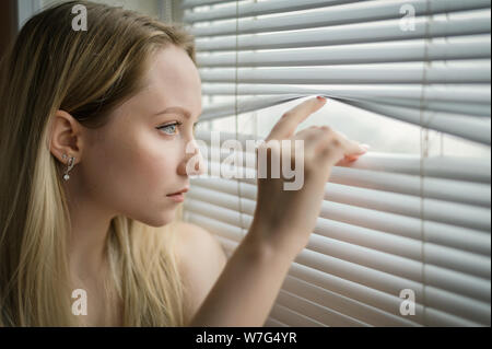 Young woman separating slats of blinds and looking through window. Stock Photo