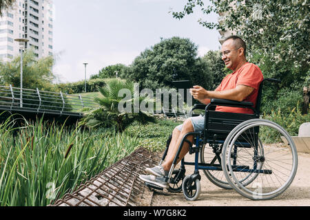 handicapped man in wheelchair having fun while working using a tablet computer at park, concept of technological and occupational integration of peopl Stock Photo