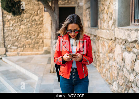 Tourist girl dressed in red jacket and sunglasses chatting with her smart phone on the street - Close-up Stock Photo