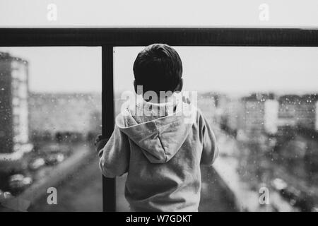Black and white shot of a child wearing a gray hoodie standing at a balcony Stock Photo