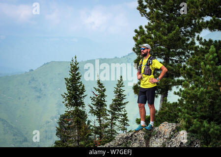 Athlete runner stands on the edge of a cliff in the mountains. Man in a yellow T-shirt and black shorts is training in outdoors. Trail running Stock Photo