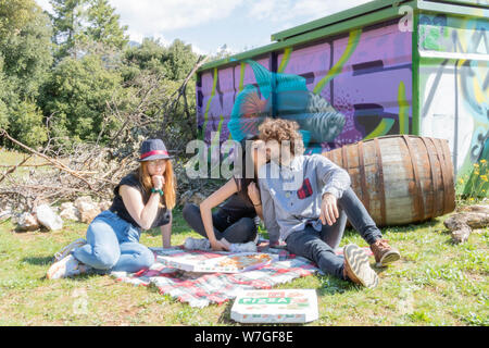 Group of young multiethnic during a picnic in the countryside to eat pizza, the excited Indian woman kisses the young Italian man in front of the Fren Stock Photo