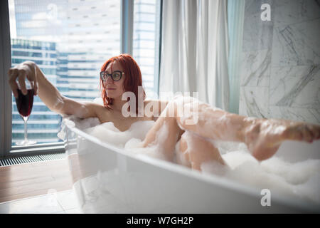 A ginger woman lying in the foam bath and looking to the side - holding a glass of red wine - a view on a modern glass buildings from the panoramic Stock Photo