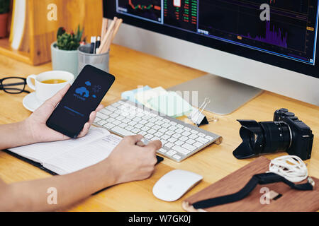 Hands of photographer using cloud application on smartphone and taking notes in planner when working at his desk Stock Photo