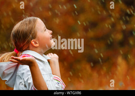 Side view of a cute charming little girl standing in the autumn park in the rain. The child reaches out and collects drops in the palm of his hand. Bl Stock Photo