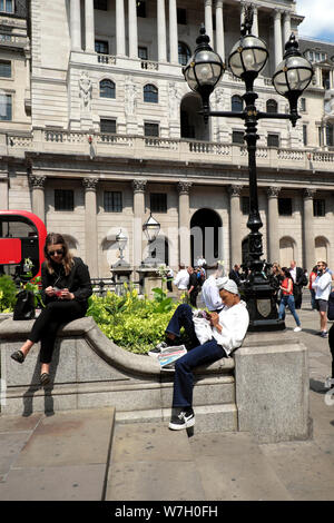 Cool young woman wearing white texting on mobile phone and pedestrians in summer sitting outside the Bank of England City of London UK  KATHY DEWITT Stock Photo