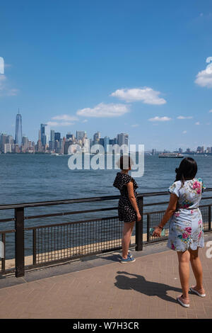 2 young woman stop to take a photo on Liberty Island with the New York City skyline in the background, including Lower Manhattan and the Freedom Tower Stock Photo