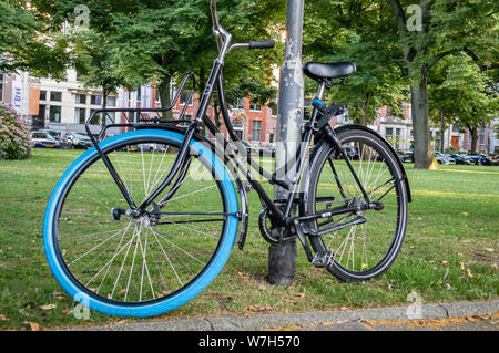 Rotterdam, Netherlands. June 29, 2019. Bike black blue colors leaned on a metal pole, buildings and parked cars background Stock Photo