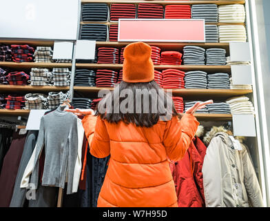 Rear view of confused woman gesturing hands to the side while looking at clothes displayed in store. A lot of warm bright colored sweaters of Stock Photo