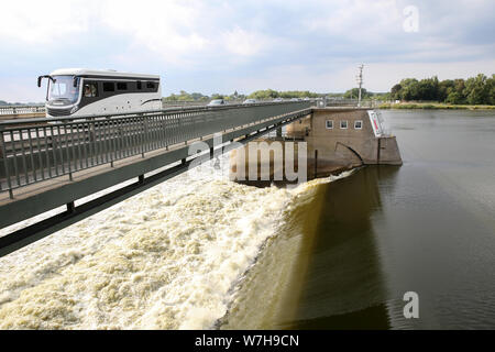 06 August 2019, Lower Saxony, Rönne: Water flows over the Elbe weir at Geesthacht, while a bus crosses the bridge. Since 02.08.2019, water had to be drained in order to reduce the water pressure on the structure and to relieve the dam. As a result, the water level dropped and shipping had to be stopped. Photo: Bodo Marks/dpa Stock Photo