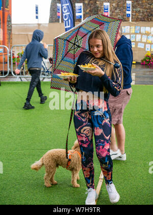 North Berwick, East Lothian, Scotland, UK, 6 August 2019. UK Weather: Torrential downpours during the festival with visitors to the seaside Fringe by the Sea in North Berwick harbour. A young pretty girl tires to balance an umbrella while holding a dog lead and takeaway chips in the rain Stock Photo