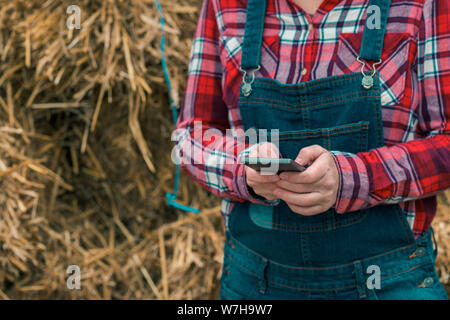 Female farmer typing sms message on mobile phone in front of the farmhouse haystack, close up of hands Stock Photo