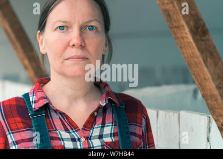 Serious female farmer posing on farm. Confident woman farm worker wearing plaid shirt and jeans overalls looking at camera, close up Stock Photo