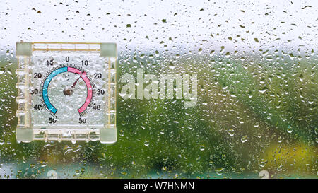 raindrops and outdoor wet thermometer on glass of home window and blurred city park on background on cool rainy autumn day Stock Photo
