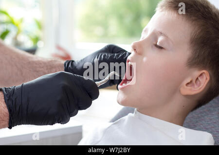 Plucking a milk tooth. The boy is sitting on a dental chair. The dentist in black rubber gloves holds the side root lever. Stock Photo