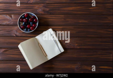 An small open notebook with pencil with bowl with cherries and blueberries on wooden desk background. Top view. Flat lay. Backdrop. Stock Photo