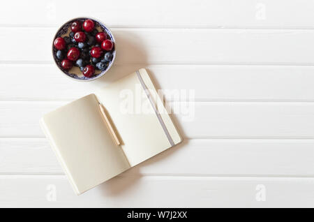 An small open notebook with pencil with bowl with cherries and blueberries on the white wooden desk background. Top view. Flat lay. Backdrop. Stock Photo