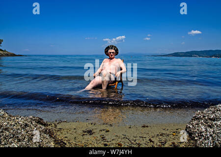 An elderly gentleman sits on a wooden chair that is on the beach in the Aegean Sea and enjoys the sun. Stock Photo