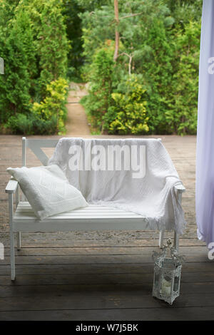White bench on the outdoor terrace with wooden floor. Behind the garden.On the bench is a blanket and pillow. A cozy place to relax Stock Photo