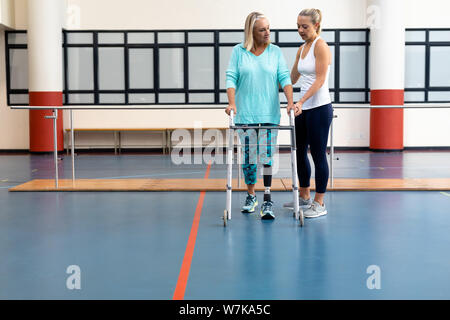 Female physiotherapist helping disabled senior woman walk with walker in sports center Stock Photo