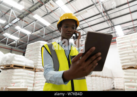 Female worker using digital tablet while talking on mobile phone in warehouse Stock Photo