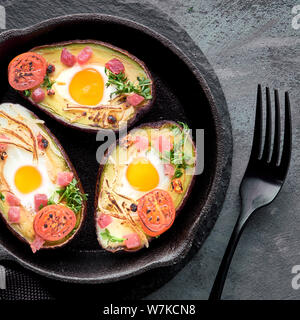 Keto diet dish: avocado boats with ham cubes, quail eggs, cheese and cherry tomatoes on iron cast skillet with a towel. Square composition, top lay on Stock Photo
