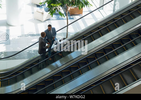Business people interacting with each other while moving upstairs on escalator in office Stock Photo