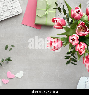 Springtime flat lay with keyboard, mouse, cards, bunch of pink tulips, eucalyptus and gift boxes on stone surface, square composition Stock Photo