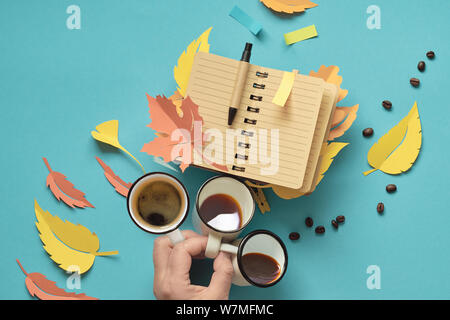 Autumn planning, creative top view flat lay on mint color paper background. Hand holding three cups of coffee and open blank notebook with autumn leav Stock Photo