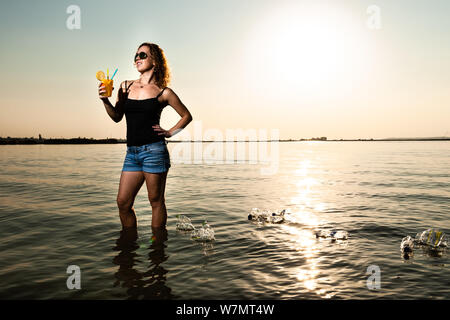 Woman in the sea holding a glass of juice with plastic straw around many plastic bottle swimming on the water. Environmental protection concept. Stock Photo