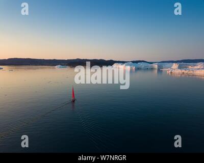 Little red sailboat cruising among floating icebergs in Disko Bay glacier during midnight sun season of polar summer. Ilulissat, Greenland  Expedition Stock Photo