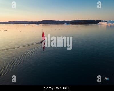 Little red sailboat cruising among floating icebergs in Disko Bay glacier during midnight sun season of polar summer. Ilulissat, Greenland  Expedition Stock Photo